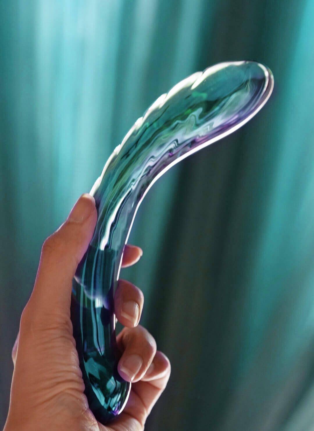 A hand holding the Kalii — rainbow purple borosilicate glass — against a teal curtain backdrop.